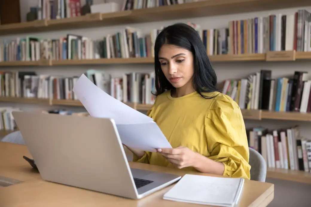 A woman reviewing documents at a laptop in a library, representing focused college planning with Opus College Prep.